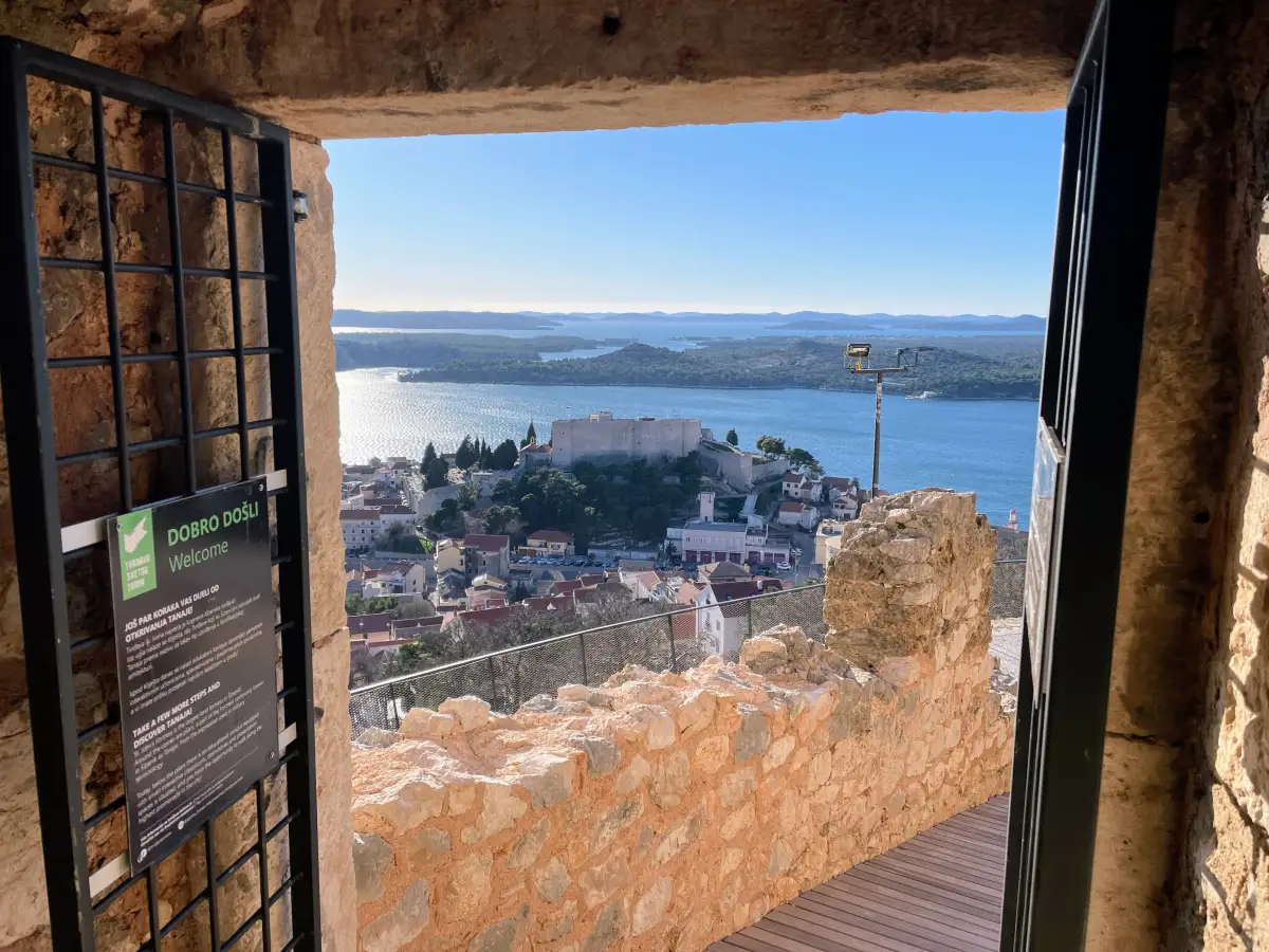 View of St. Michael's Fortress seen through a gate from St. John's Fortress in Šibenik, showcasing the historic architecture against a backdrop of the Adriatic Sea and distant islands, under a clear blue sky.