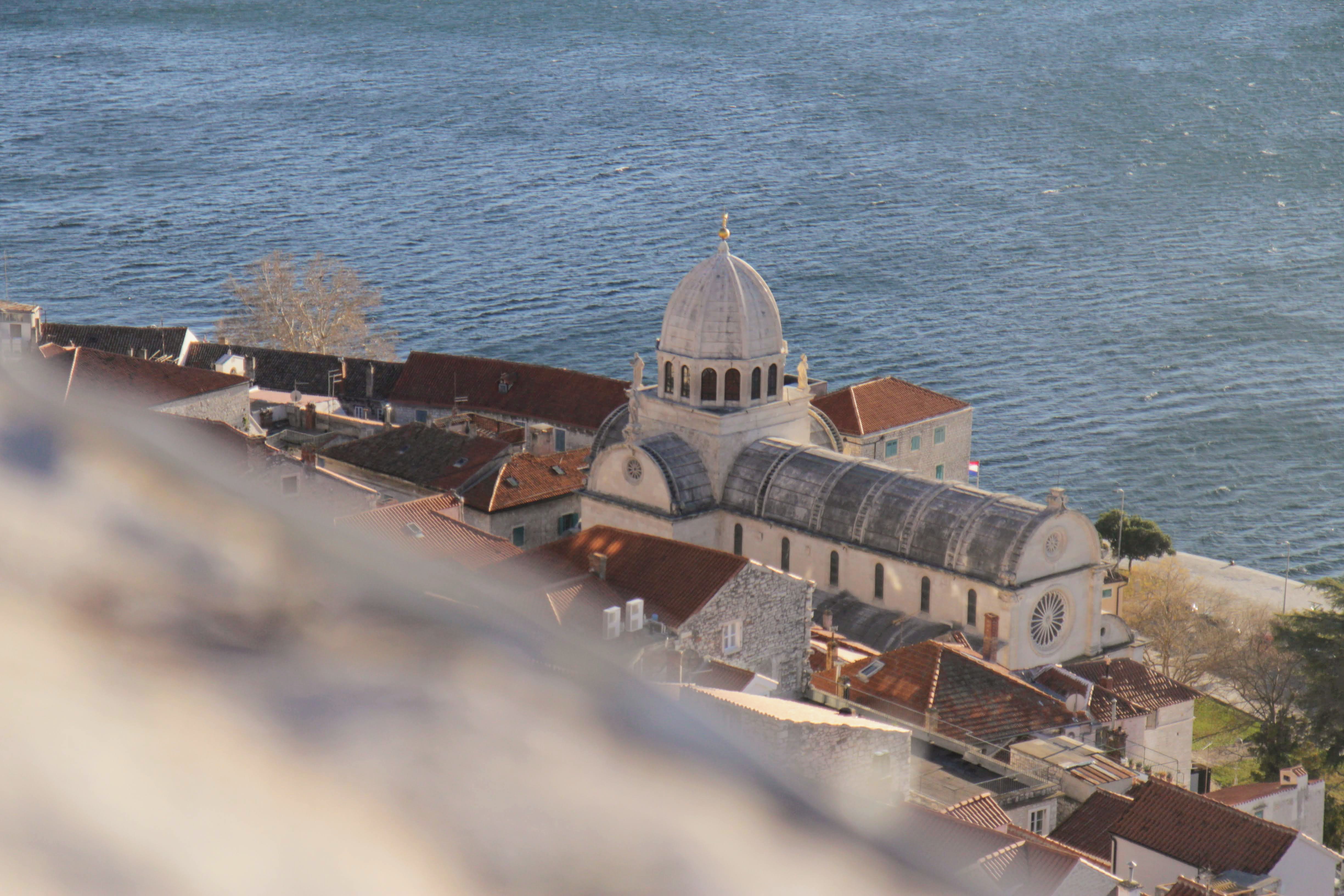 Aerial view of St. James Cathedral in Šibenik, Croatia, with its white dome rising above terracotta roofs. The Adriatic Sea stretches out behind the historic coastal town.
