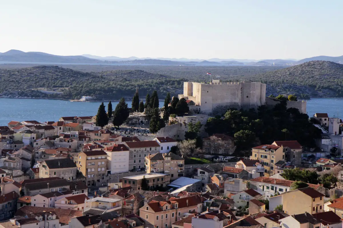 The imposing St. Michael's Fortress stands above the terracotta rooftops of Šibenik's old town, with the serene Adriatic Sea and islands in the background.