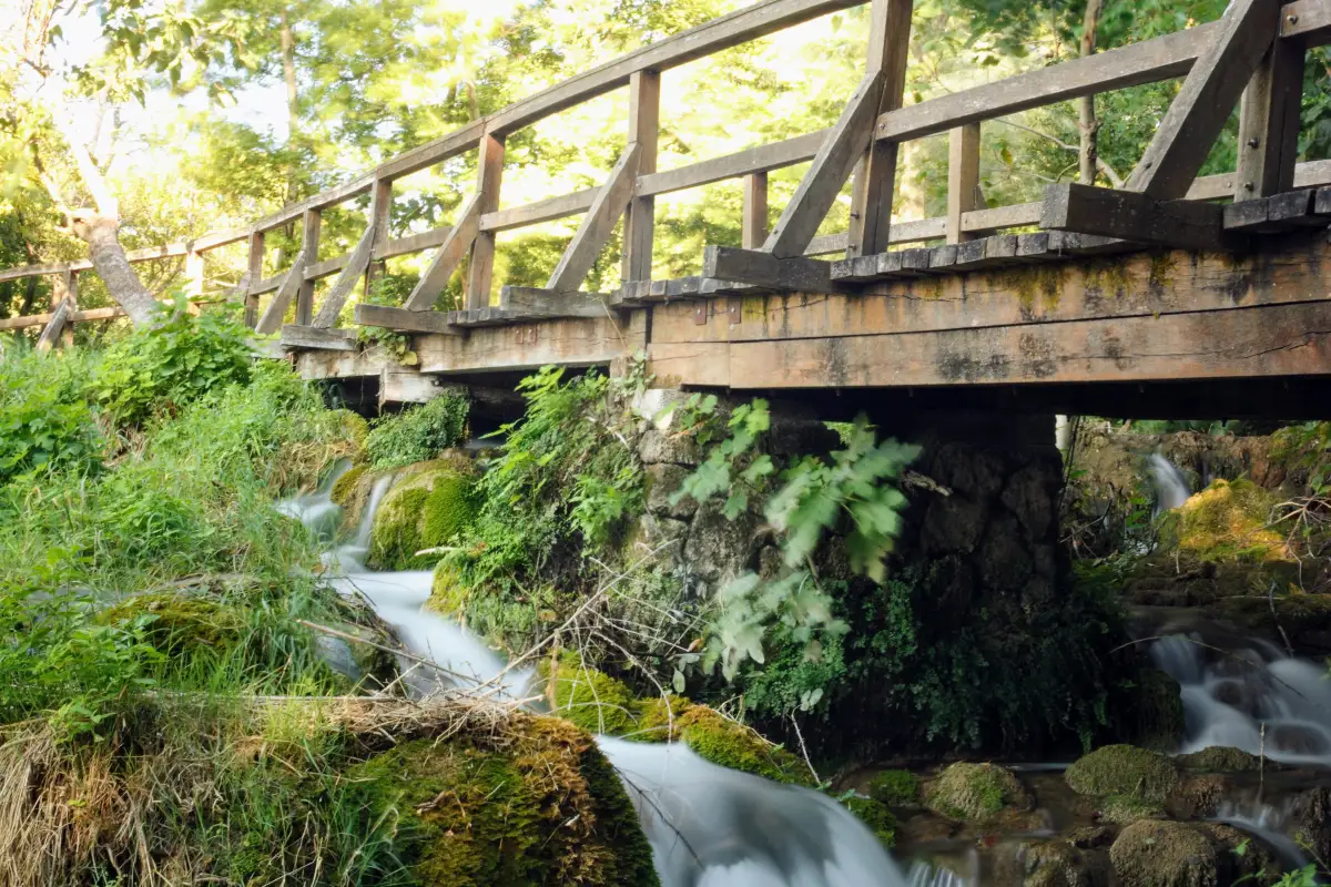 Rustic wooden bridge crossing over a small, clear stream with cascades, surrounded by lush vegetation in Krka National Park.