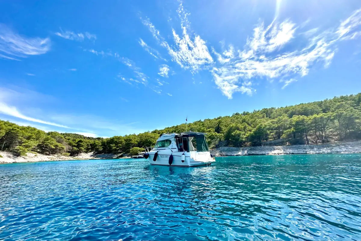A white boat floating on the clear blue waters of a calm sea, with a backdrop of a lush green coastline under a bright blue sky with wispy clouds.