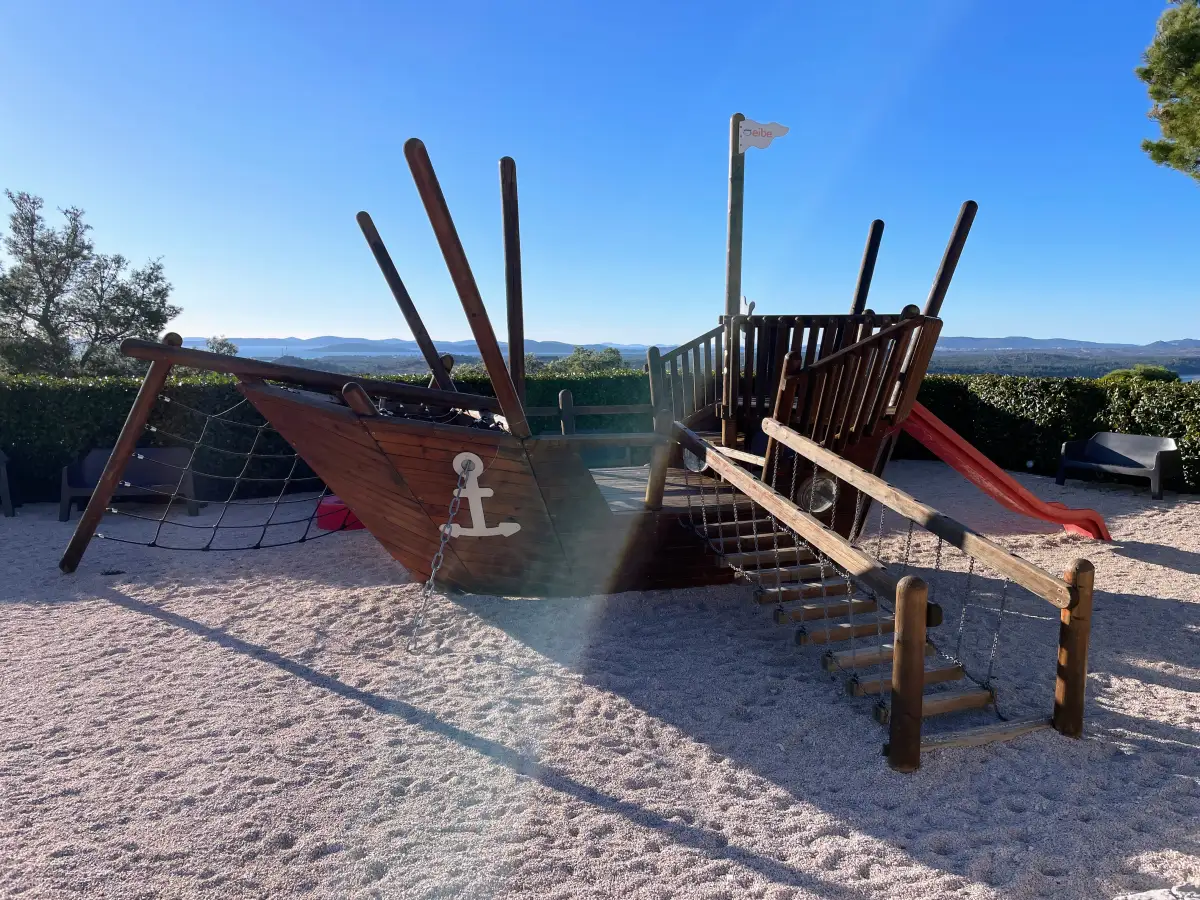 Children's playground with a wooden pirate ship structure on a sandpit on the Barone fortress, including a slide and climbing net, with a panoramic backdrop of the Dalmatian landscape under a clear blue sky.