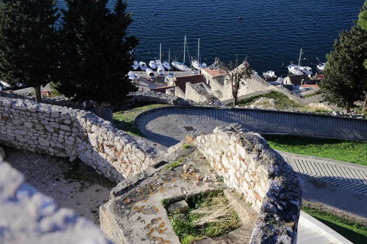 Elevated view of a serpentine walkway descending to a marina with moored boats in Šibenik, as seen from St. Michael's Fortress. It shows the double walls, often called the path of salvation