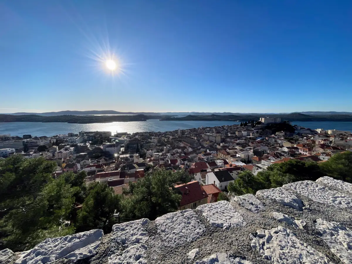Panoramic view of Šibenik and its coastline, with the sun casting a glittering reflection on the Adriatic Sea, as seen from the heights of a fortress.