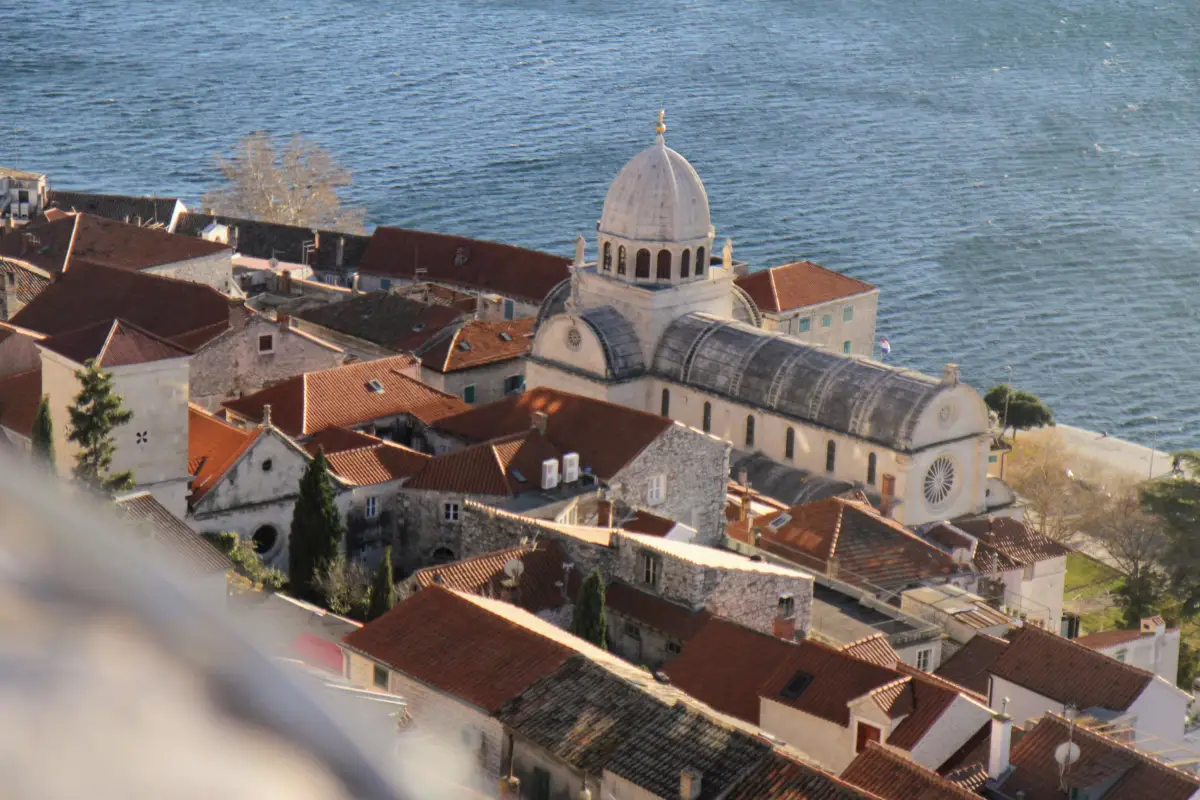 levated view of the iconic Šibenik Cathedral with its white dome, nestled among the red-tiled roofs of the old town, adjacent to the Adriatic Sea.