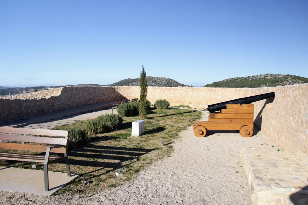 Historical St. John fortress wall with a traditional cannon on a wooden carriage overlooking a serene landscape with Mediterranean flora under a bright blue sky.