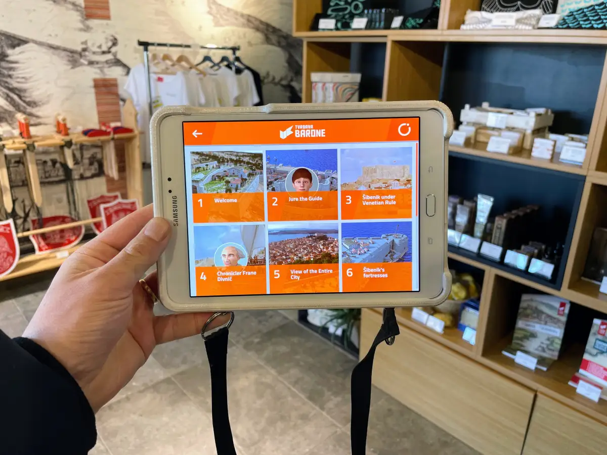 Hand holding a tablet with an interactive tour guide application showing various historical points of interest at Barone Fortress, with souvenir shop shelves in the background.