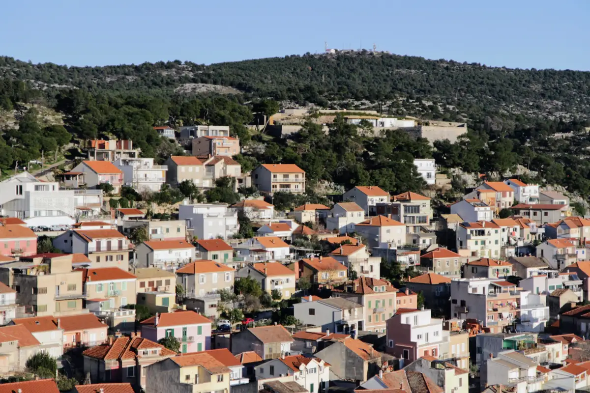 Close-up view of Šibenik's residential area with multi-colored houses and red-tiled roofs ascending towards a historic fortress on a hill, under the bright blue sky, highlighting the town's urban and historical landscape.
