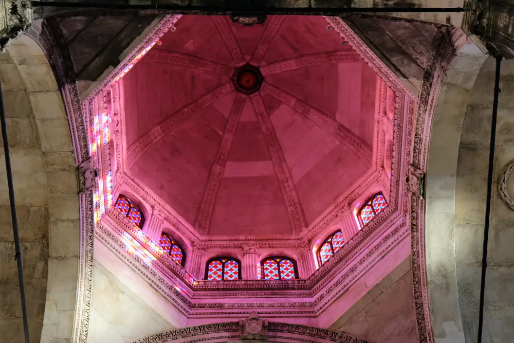 interior view of the dome of St. James Cathedral in Šibenik, Croatia. The dome's intricate design features Gothic and Renaissance architectural elements, illuminated by colorful stained glass windows that cast a pinkish hue across the stonework.