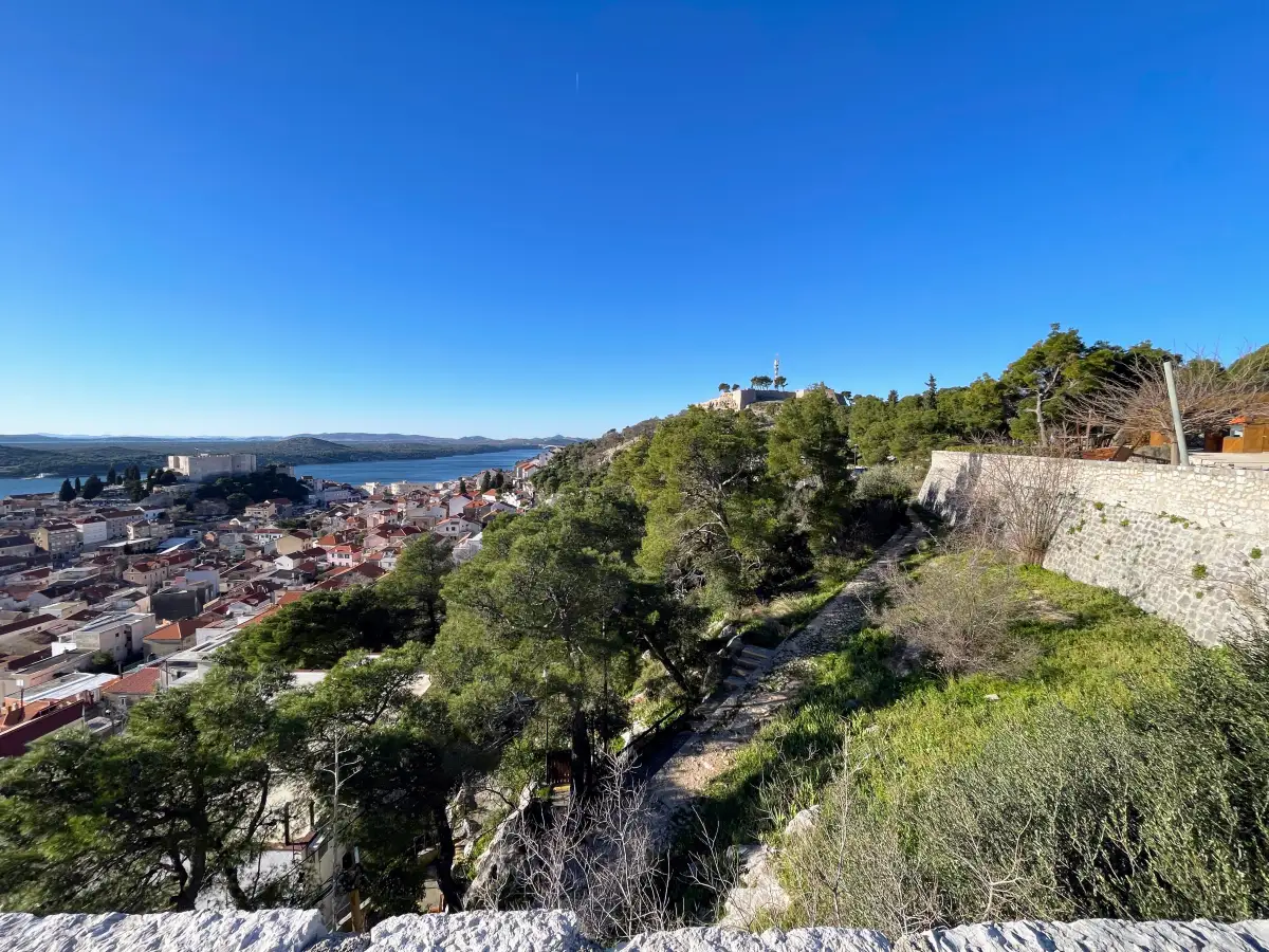 View from Šibenik panoramic fortress across the historic cityscape with roofs of red-tiles framed by green Mediterranean vegetation and the Adriatic Sea in the distance beyond, beneath a clear blue sky.