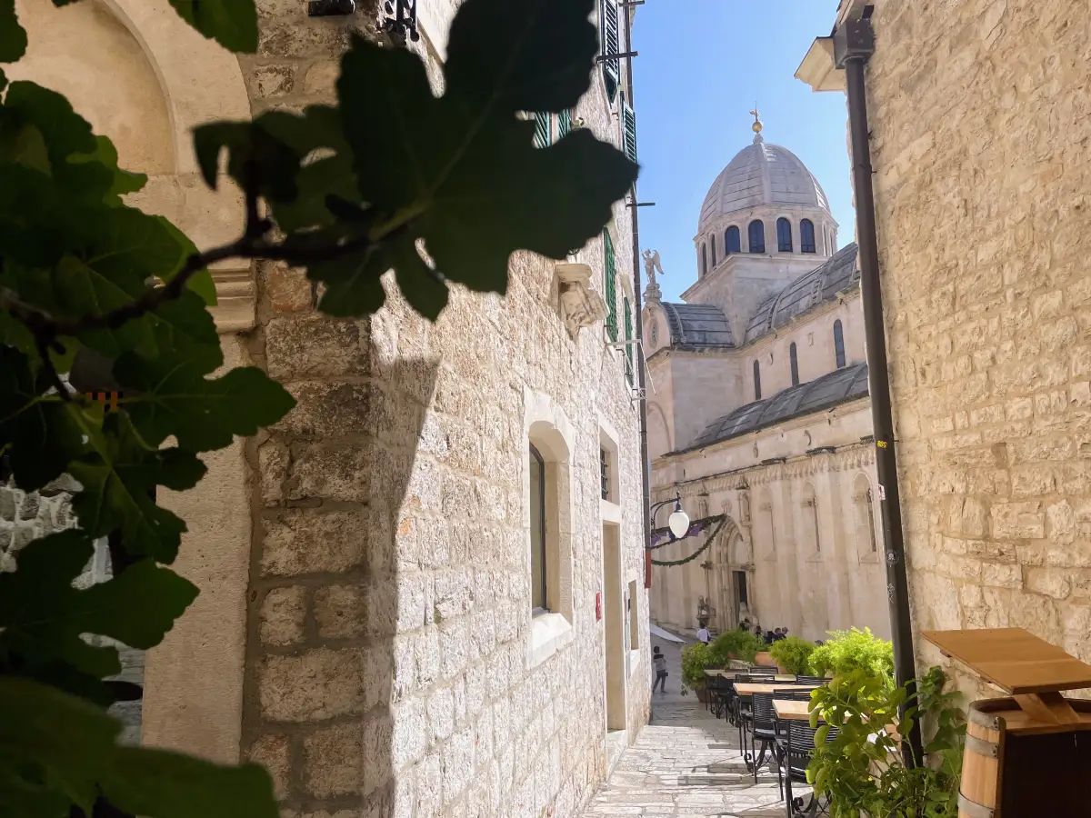 A narrow alley leading to the Cathedral of St. James in Šibenik, framed by stone buildings and green foliage, with a glimpse of the cathedral's dome against a clear blue sky.