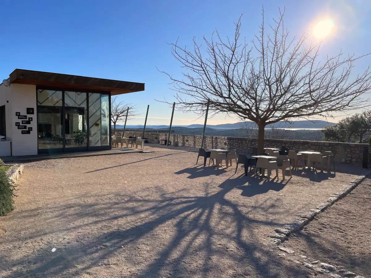 Café at Barone Fortress in Šibenik with outdoor tables, a leafless tree casting shadows on the ground, and a view of distant hills under a clear sky.
