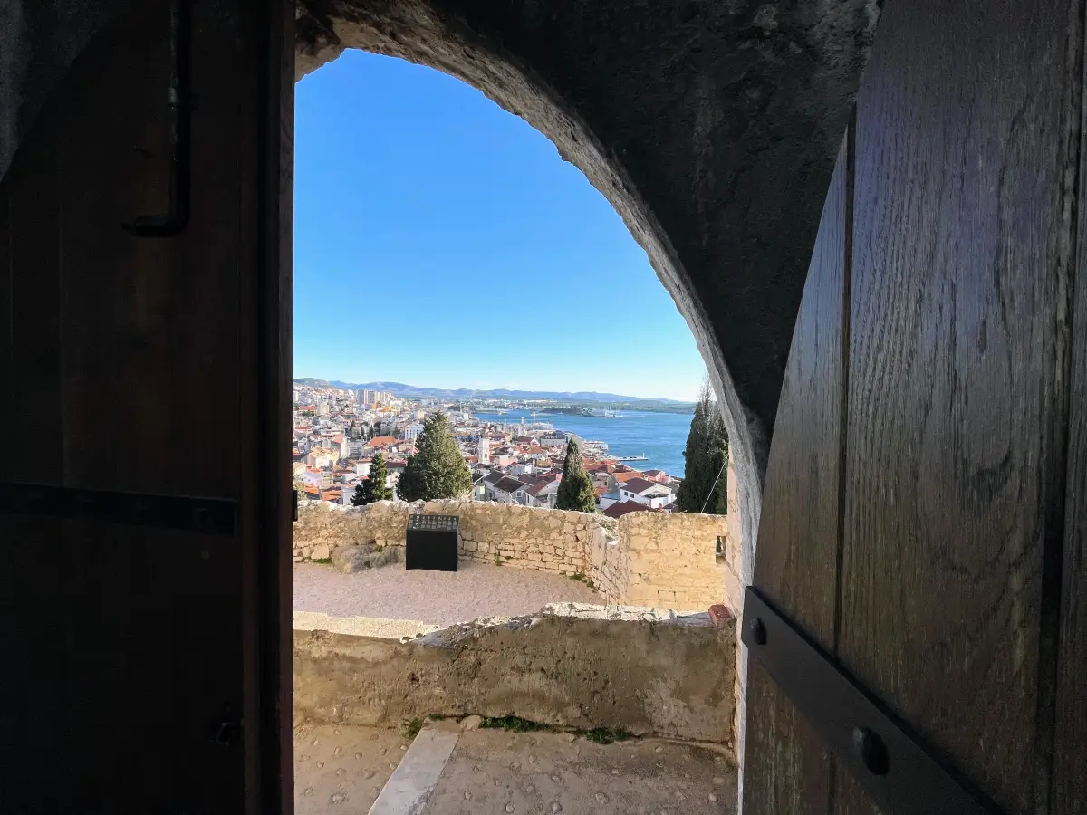 View of Šibenik's old town and the Adriatic Sea through an arched gateway at St. Michael's Fortress, framed by a wooden door.