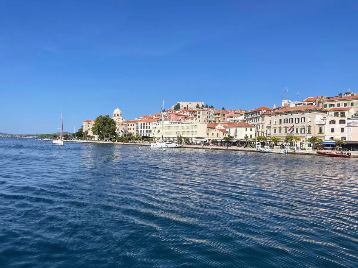 Panoramic view of Šibenik's waterfront promenade, showcasing the Cathedral of St. James and traditional Mediterranean architecture along the clear blue waters of the Adriatic Sea on a sunny day.
