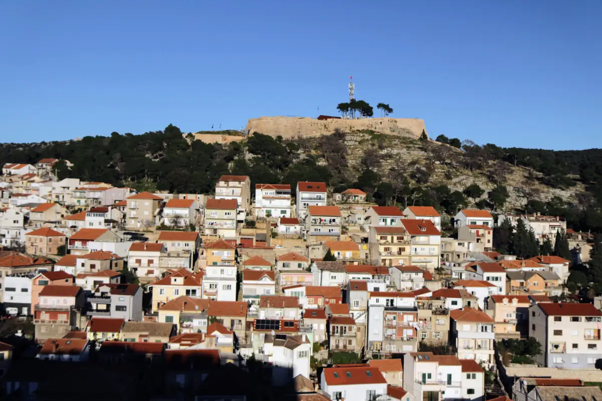 A dense array of traditional Mediterranean houses in Šibenik ascending toward a hilltop fortress under a bright blue sky, demonstrating the historical overlay of the city's architecture and its natural landscape.