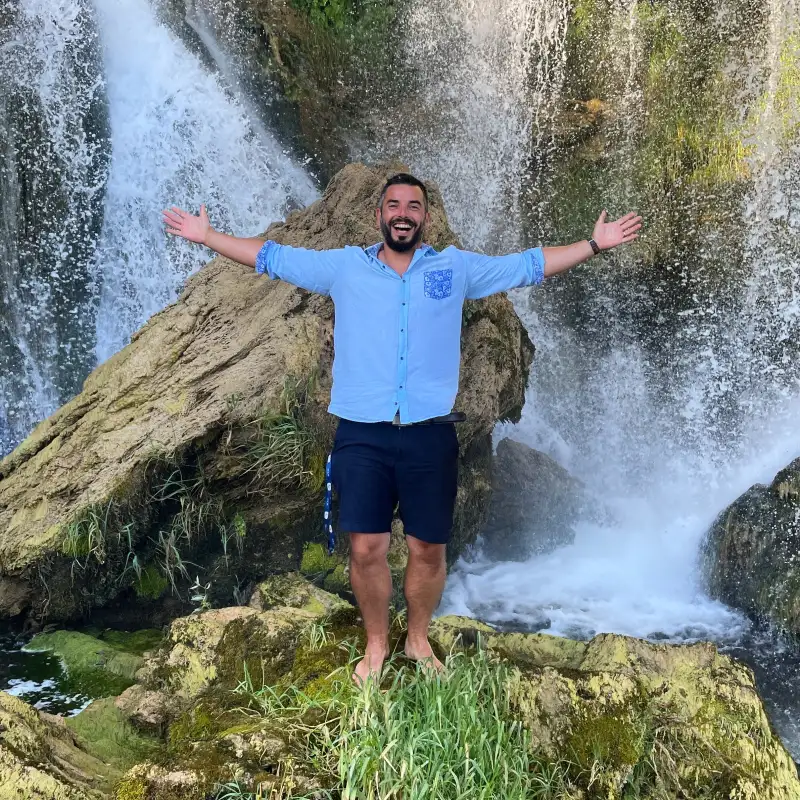 A man with a wide smile extends his arms wide open, standing in front of a cascading waterfall. He's dressed in a light blue shirt and navy shorts, barefoot on the rocky edge, conveying a sense of welcoming and the grandeur of nature around him.