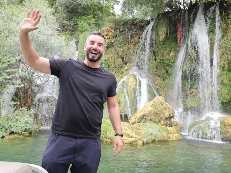A man with a friendly smile waves to the camera, standing in front of a lush waterfall. He wears a casual black t-shirt and sports a short beard and trimmed hair, exuding a sense of joy and adventure in a natural, scenic setting