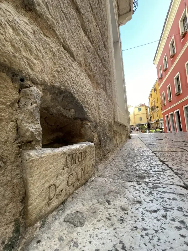 Perspective view from the ground of a weathered stone gutter with inscriptions, leading to a carved hole in the pavement of a narrow alley with colorful buildings and a clear sky above.