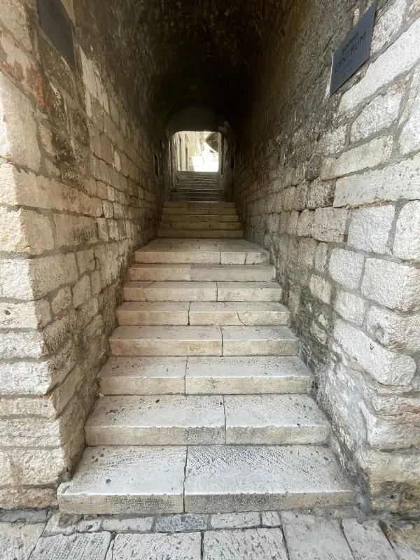 A view looking up a narrow stone staircase within a tunnel-like passageway, with sunlight filtering through the exit at the top, illuminating the textured walls and worn steps, indicative of an old, historical structure.